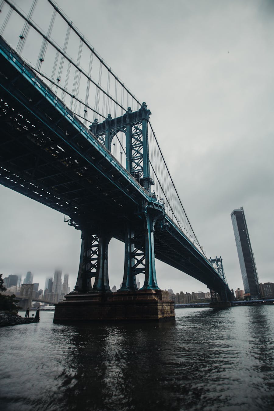 View from below of a large suspension bridge over a river with a city skyline and a tall leaning building in the background under a cloudy sky.