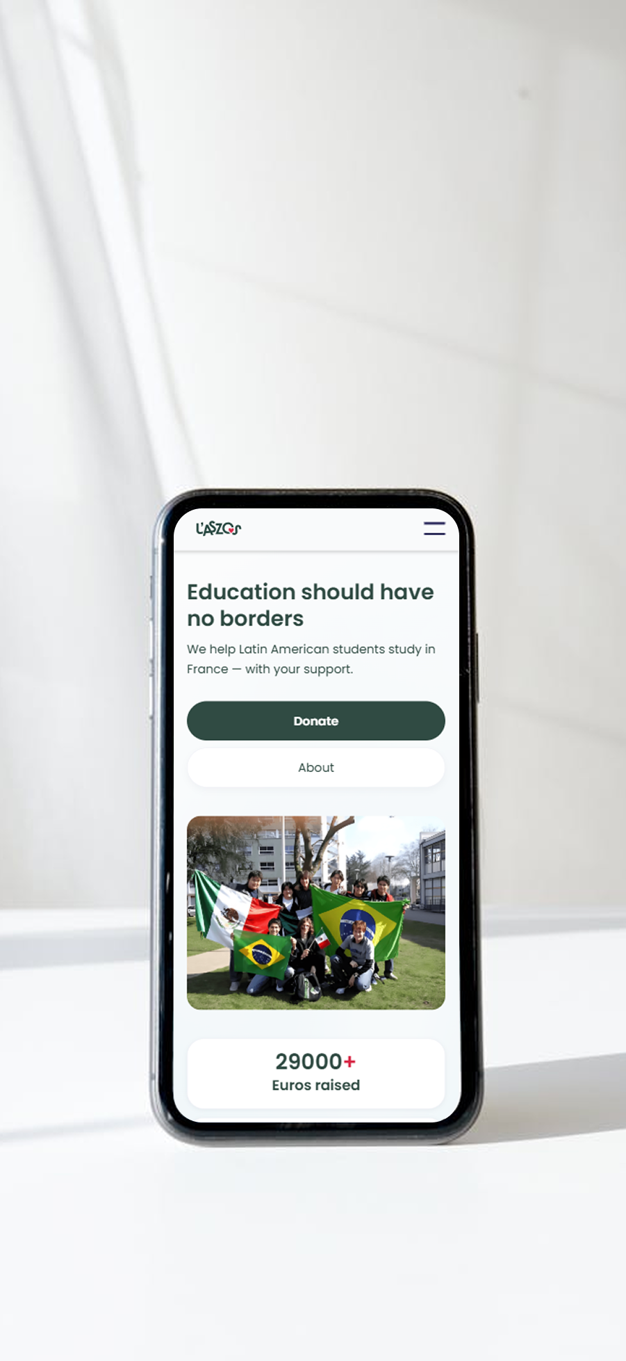 Mobile phone displaying a website promoting support for Latin American students studying in France, showing a group photo with Mexican and Brazilian flags and a note about 29,000 euros raised.